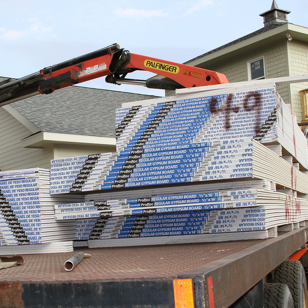 A forklift is lifting stacks of gypsum board on a truck outside a construction site, with a house partially visible in the background.