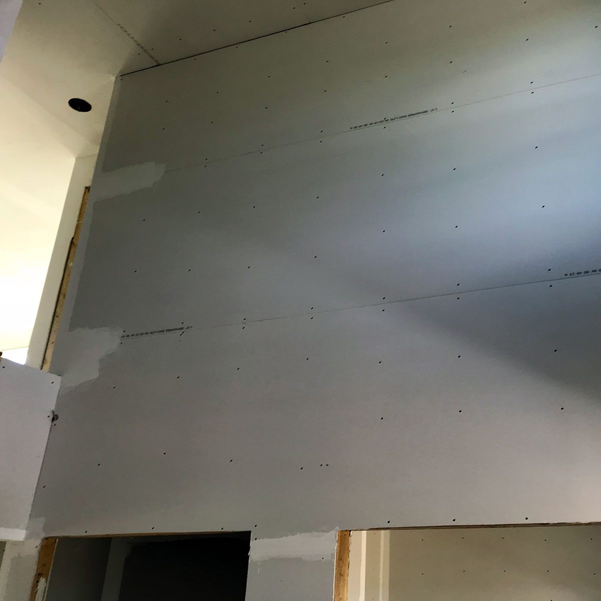 A drywall section is mounted on a wall, showing a flat surface with dimples from screws and wet joint compound, in a partially finished indoor space.