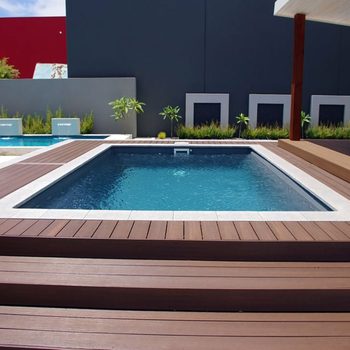 A rectangular swimming pool with clear water is surrounded by wooden decking and small green plants, set against a modern gray wall and colorful backdrop.