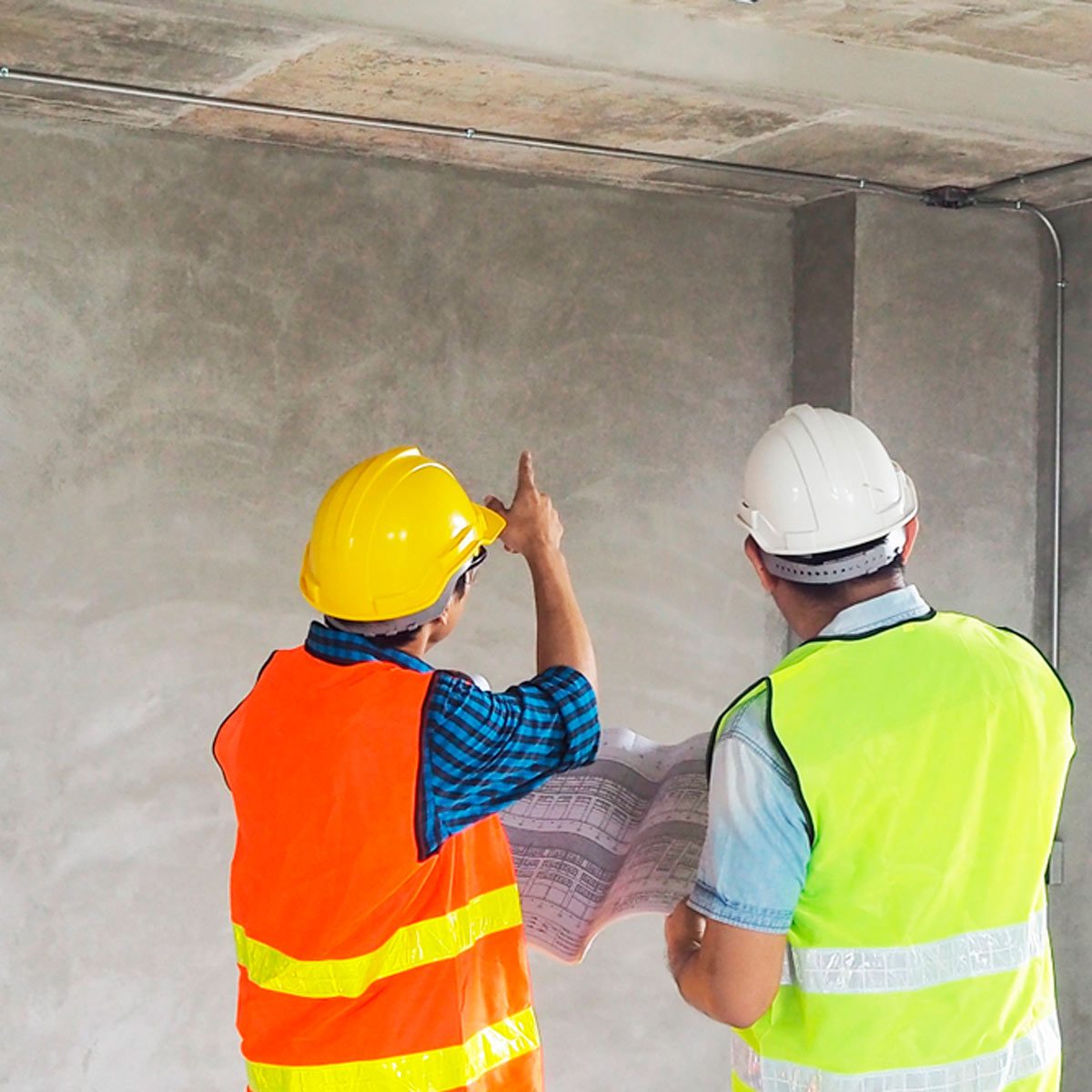 Two construction workers, one pointing and discussing plans, stand in a concrete room, wearing safety helmets and bright vests, reviewing a blueprint.