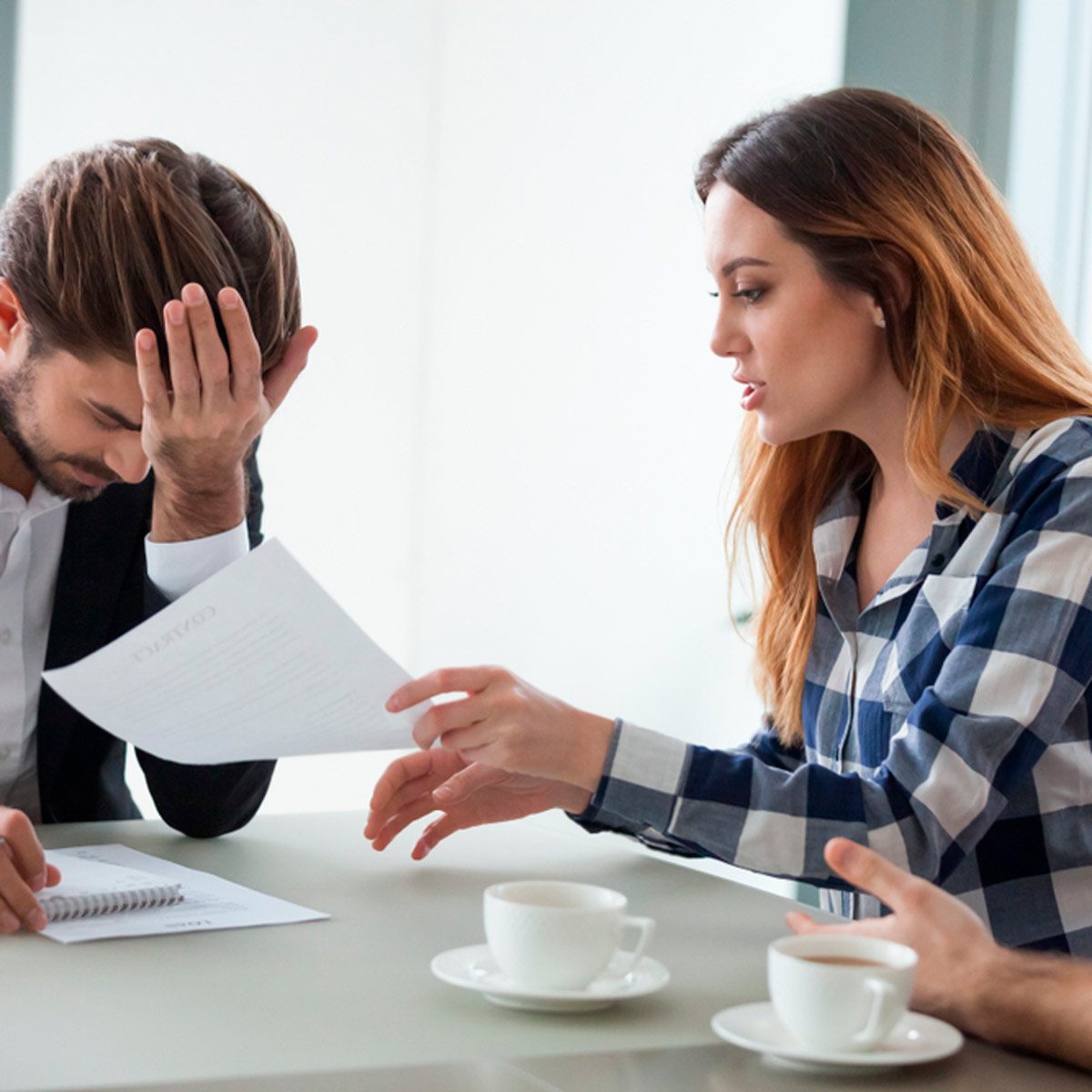 A woman discusses a document with a man, who has his head in his hand, at a table with coffee cups in a bright room.