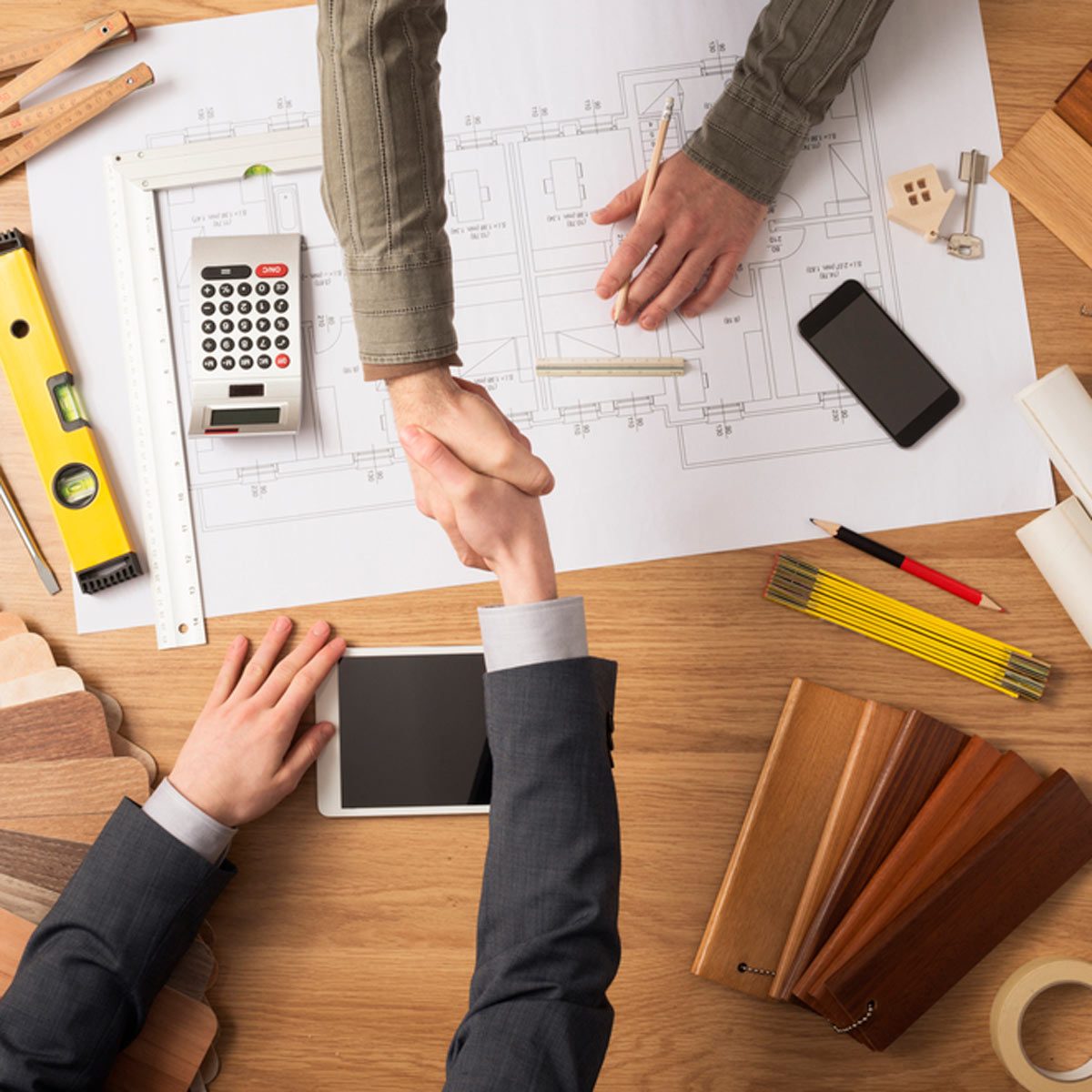 Two hands are shaking above a blueprint on a wooden table, surrounded by tools, a calculator, a smartphone, and color samples.