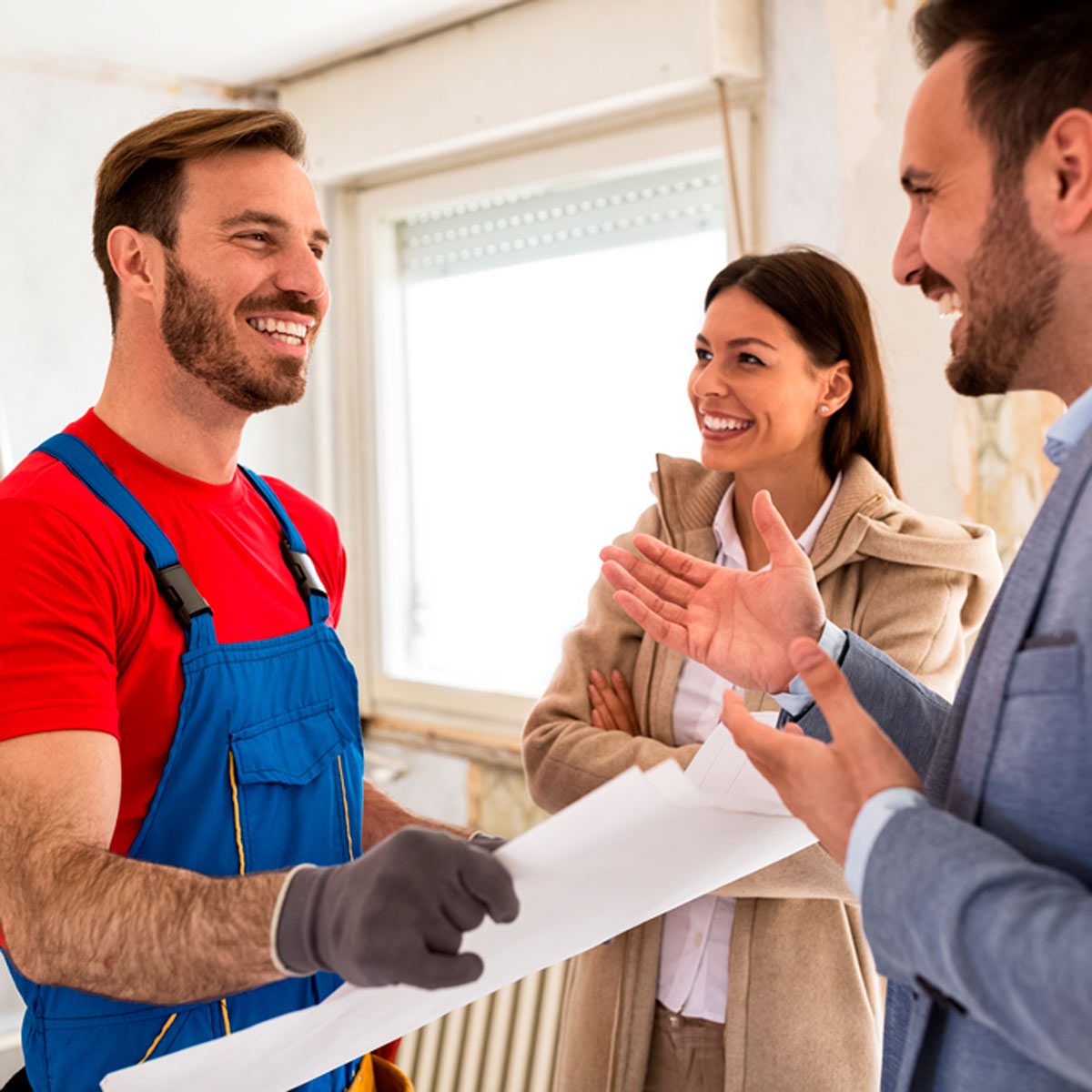 A construction worker smiles while discussing blueprints with a couple in a partially renovated room, surrounded by bare walls and natural light.