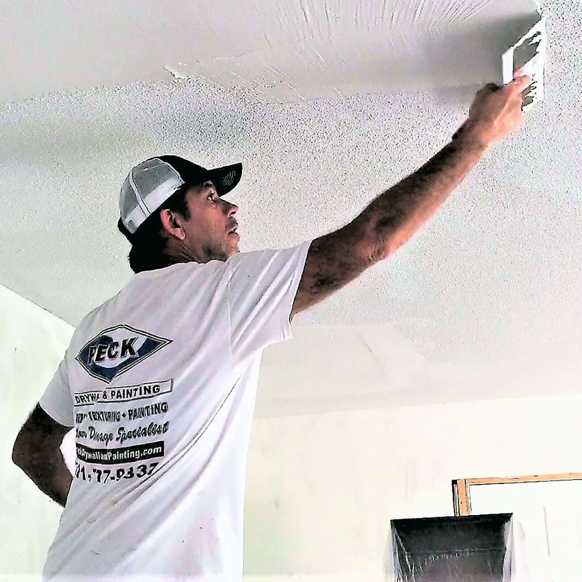 A man with a cap applies texture to a ceiling using a spatula, standing in a partially painted room with an open window.