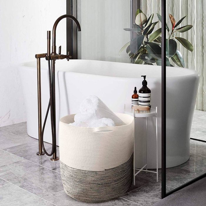A woven basket holds white towels beside a freestanding bathtub. A faucet and skincare products are nearby, set in a modern, light-toned bathroom.