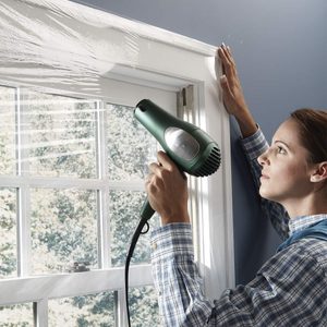 A woman uses a green hairdryer to warm plastic film over a window, preparing it for insulation in a well-lit, indoor setting.