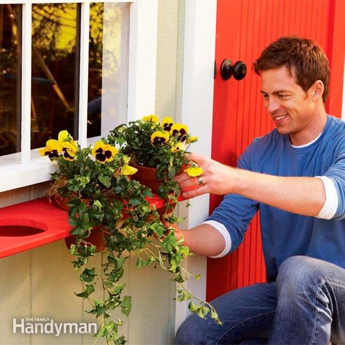 A man places potted yellow flowers on a bright red window sill, enhancing a light-colored house’s exterior while enjoying a sunny day.