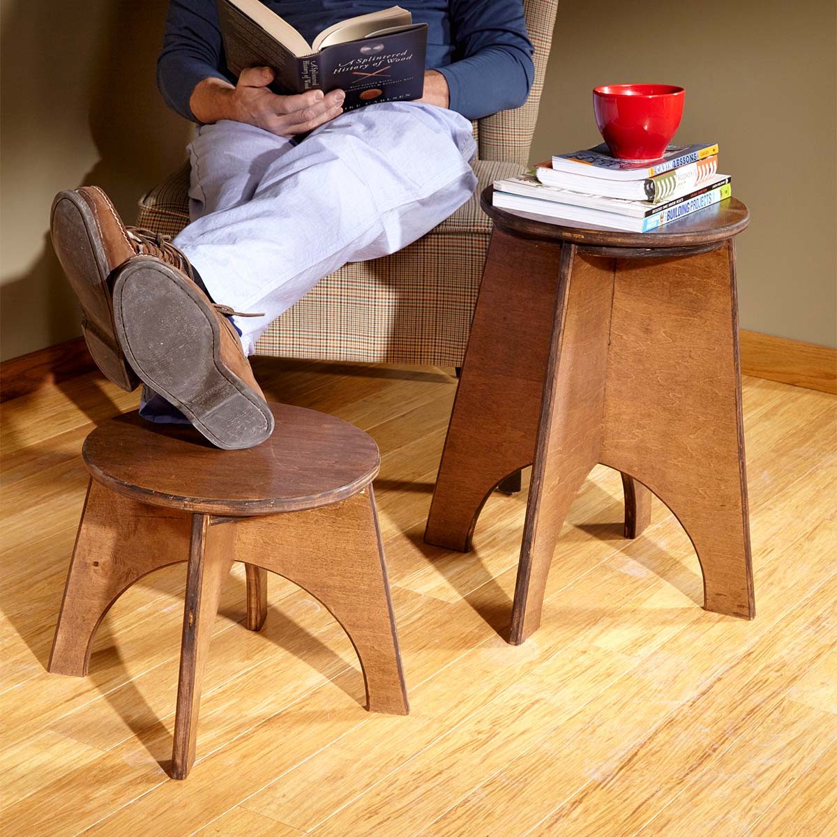 A person reads while seated in a chair, resting feet on a small wooden stool, with a larger stool nearby, holding books and a red cup.