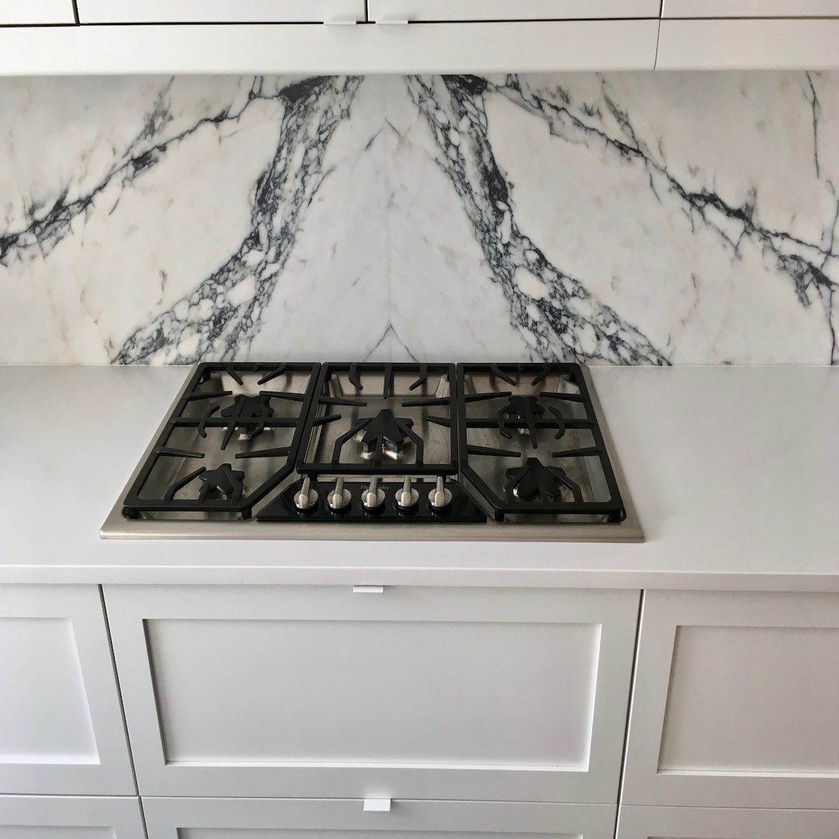 A gas cooktop with five burners sits on a white countertop, against a marble backsplash featuring black veining, in a modern kitchen setting.