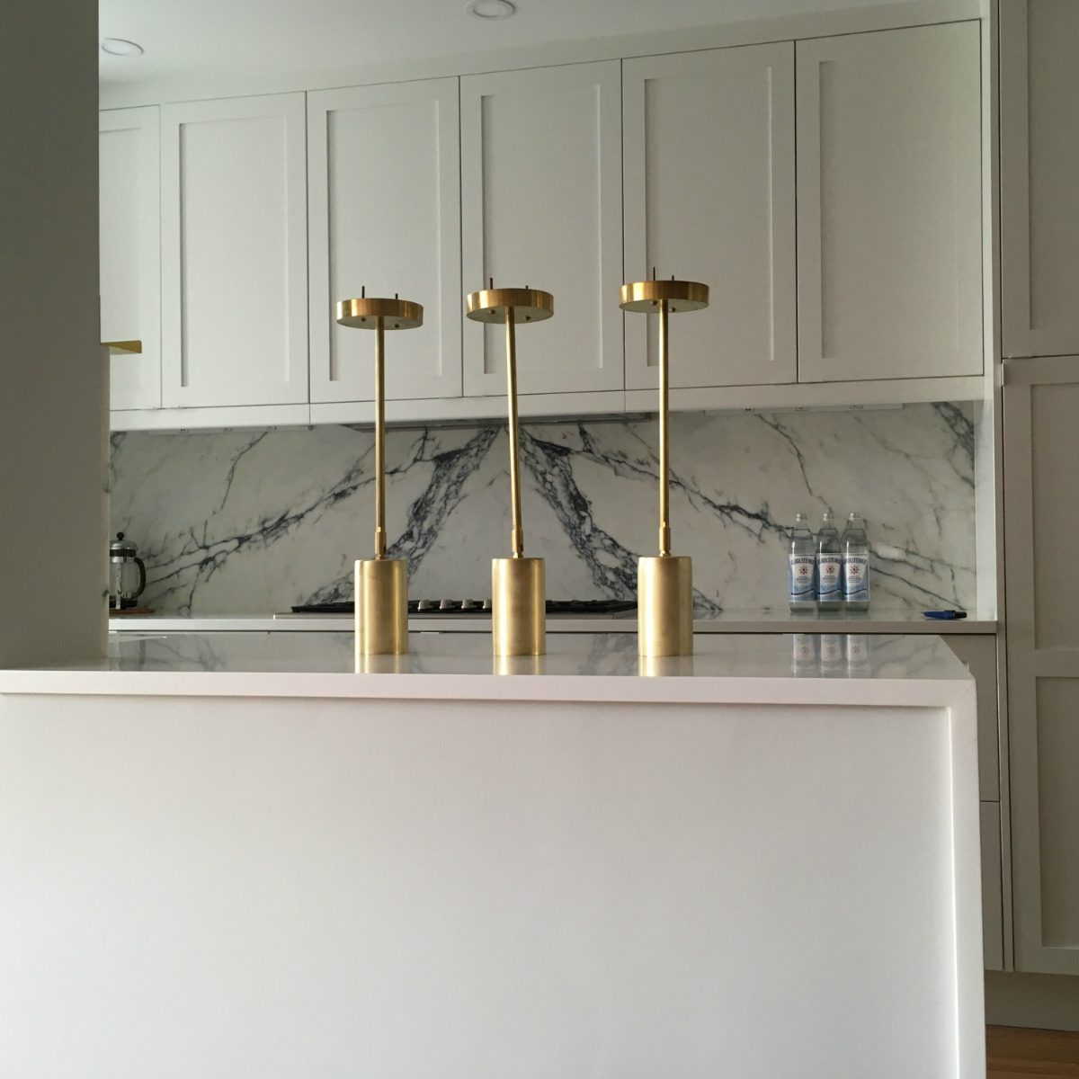 Three brass lamps stand on a kitchen island, illuminated in a modern setting, with marble backsplash and cabinetry in the background, creating an elegant atmosphere.