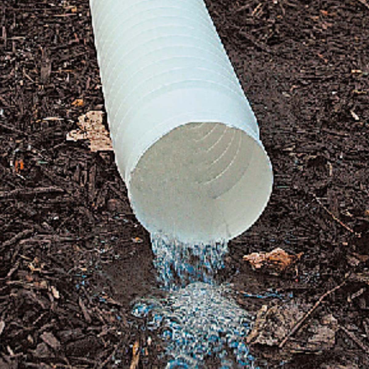 A white drainage pipe discharges water onto dark, mulched soil, creating a small pool as leaves and debris surround the area.