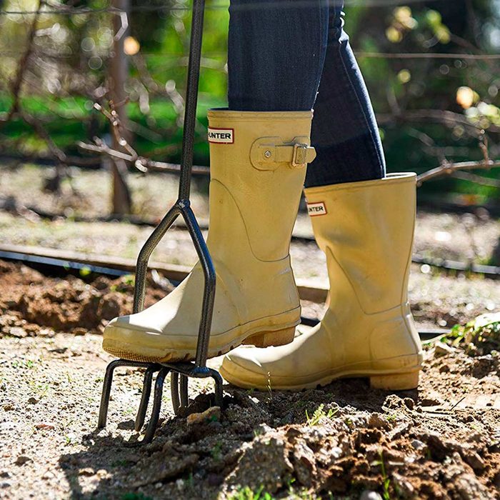 Yellow rubber boots stand on soil while a person uses a garden fork to dig in a sunlit outdoor garden surrounded by green plants.