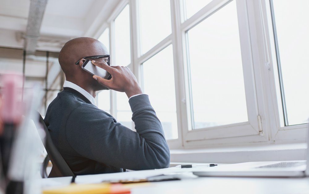 A man sitting at a desk is talking on his phone while looking out of a large window in a bright, modern office space.