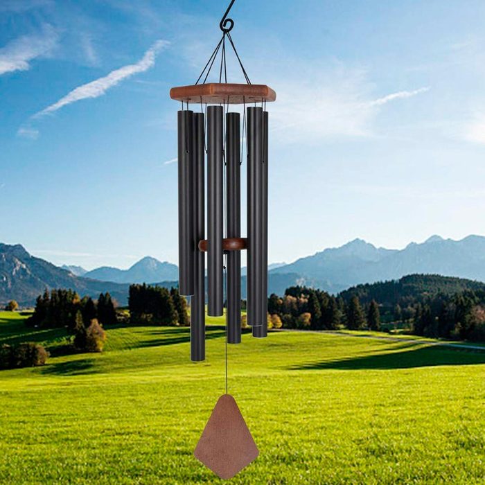 A wind chime hangs, swaying gently in the breeze, in a picturesque landscape of rolling green fields and distant mountains under a clear blue sky.