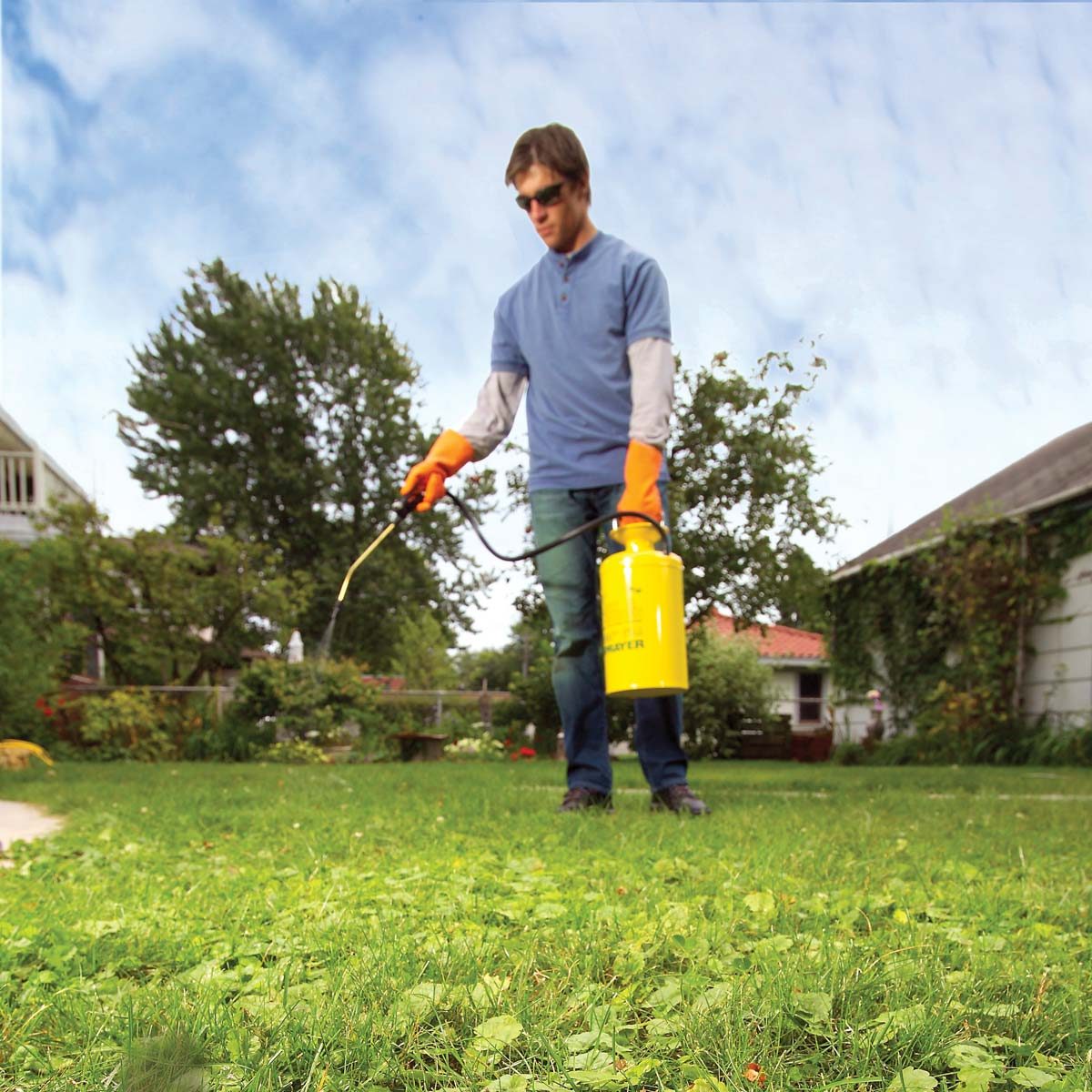 A man in orange gloves sprays chemicals on the grass with a yellow sprayer in a yard surrounded by trees and a house under a blue sky.