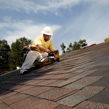 A worker uses a nail gun to install shingles on a sloped roof under a blue sky, surrounded by trees and rooftops.