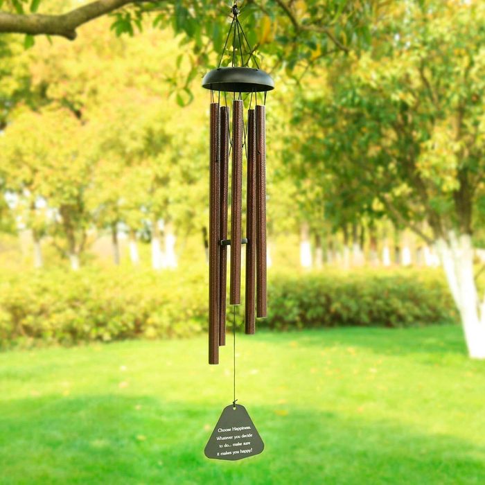 Wind chimes, hanging and swaying, produce sound in a lush green outdoor setting, with trees and a grassy area nearby.