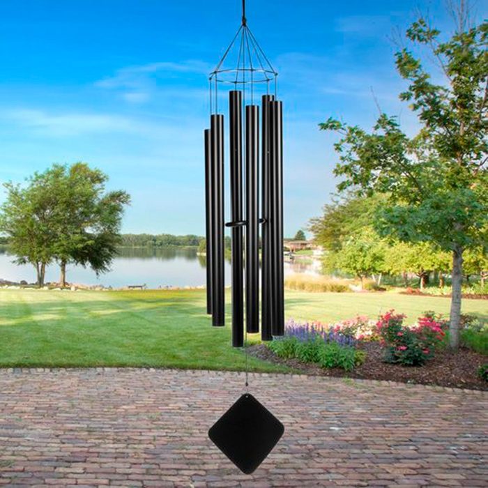 A set of black wind chimes hangs from a frame, gently swaying in the breeze above a scenic lakeside garden filled with colorful flowers.