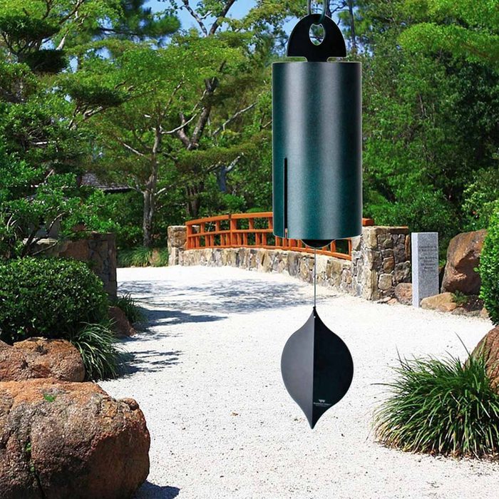 A metal wind chime hangs above a gravel path, gently swaying in the breeze, surrounded by greenery and an arched wooden bridge in the background.