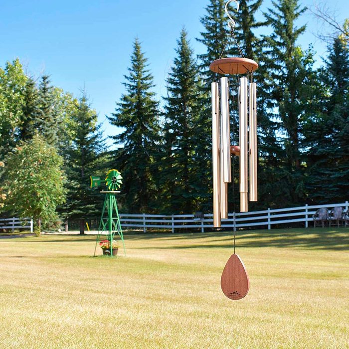A wind chime hangs gently, producing sound as the breeze moves through its tubes, set in a grassy yard with tall trees and a distant windmill.
