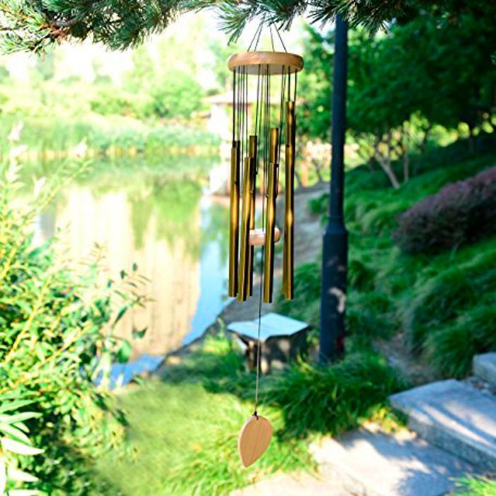 A wind chime hangs from a branch, producing sound as it sways in the breeze above a peaceful pond surrounded by greenery and pathways.