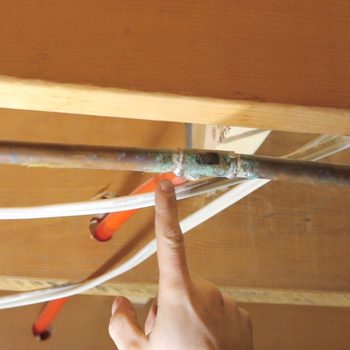 A hand points at a copper pipe connected to white and orange pipes, situated beneath a wooden surface in a presumably utility area.