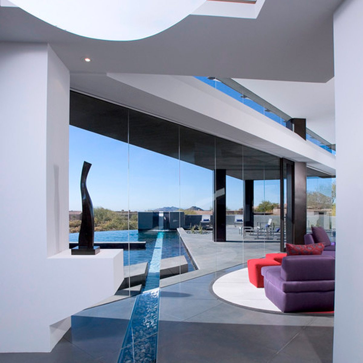 A modern living space features purple and red seating, a sleek sculpture, and large glass windows overlooking a serene pool and distant landscape.
