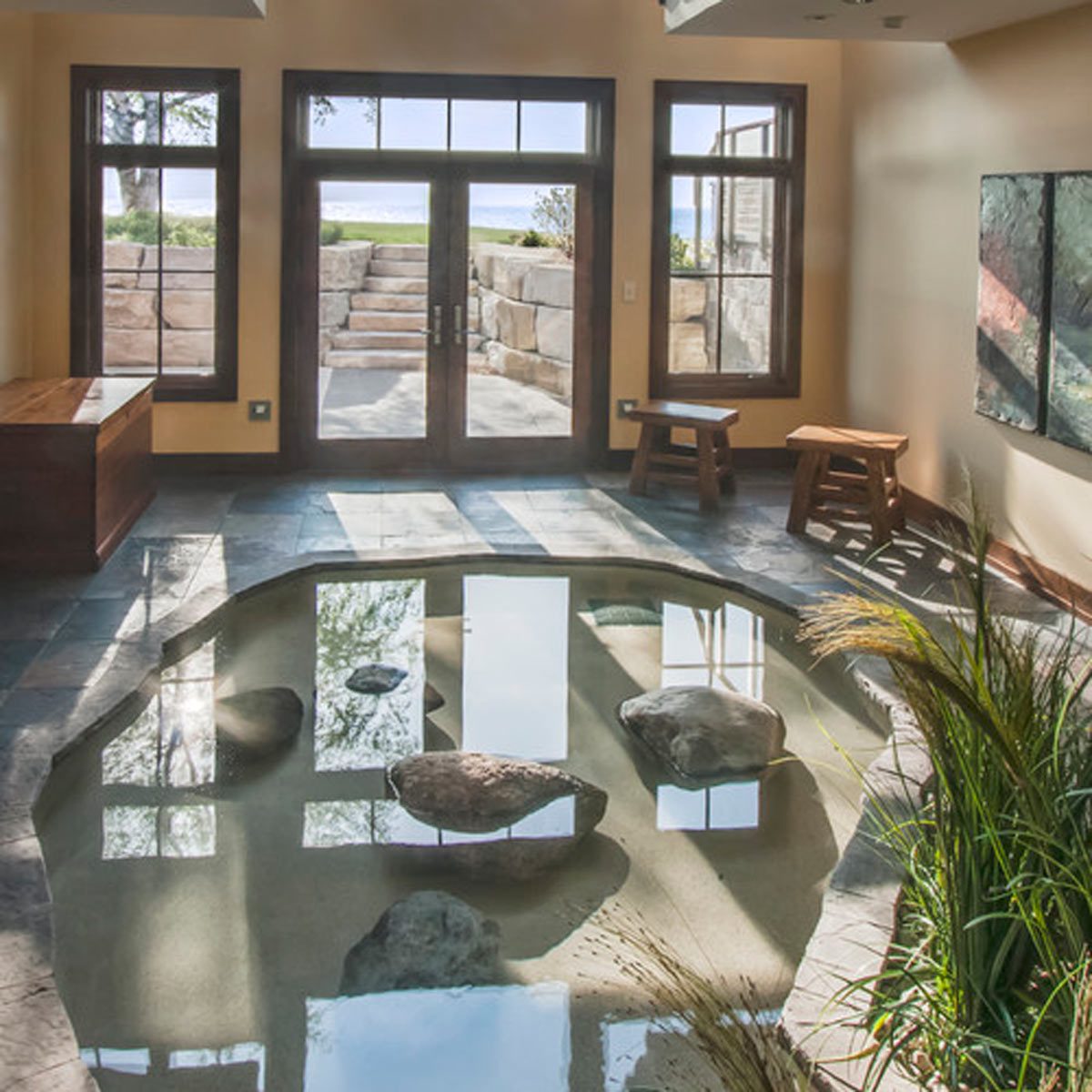 A shallow pond with rocks reflects sunlight inside a spacious, brightly lit room featuring wooden furniture and large windows overlooking a stone pathway and outdoor greenery.