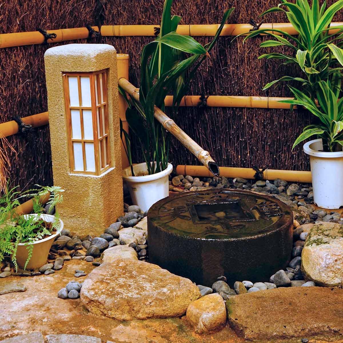 A stone basin sits surrounded by pebbles and plants, with a bamboo spout directing water into it, set against a textured bamboo fence.