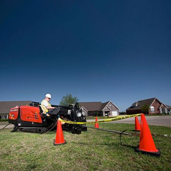 A construction worker operates a machine on grass, surrounded by orange cones and caution tape, with residential houses in the background under a clear blue sky.