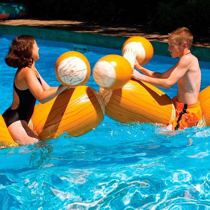 Two children play in a swimming pool, using large inflatable drumsticks as they engage in a friendly water fight. Sunlight reflects off the water.