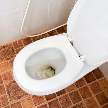 A white toilet is positioned on a tiled floor, partially filled with crumpled paper. The surrounding area features light-colored walls and plumbing connections.