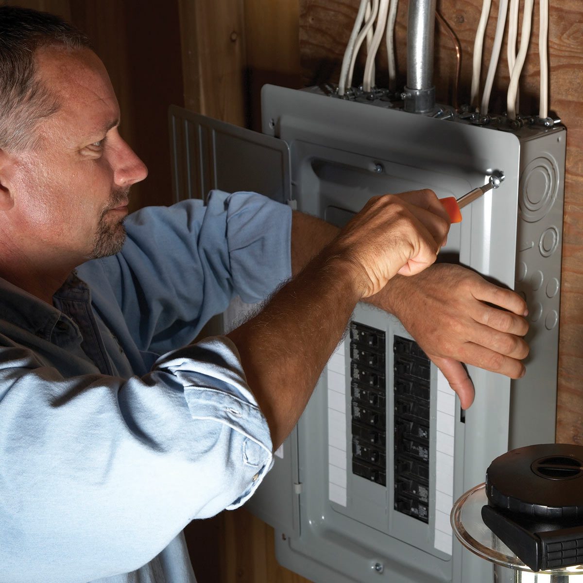 A man is tightening a screw on an electrical panel inside a wooden structure, using a screwdriver under artificial lighting.