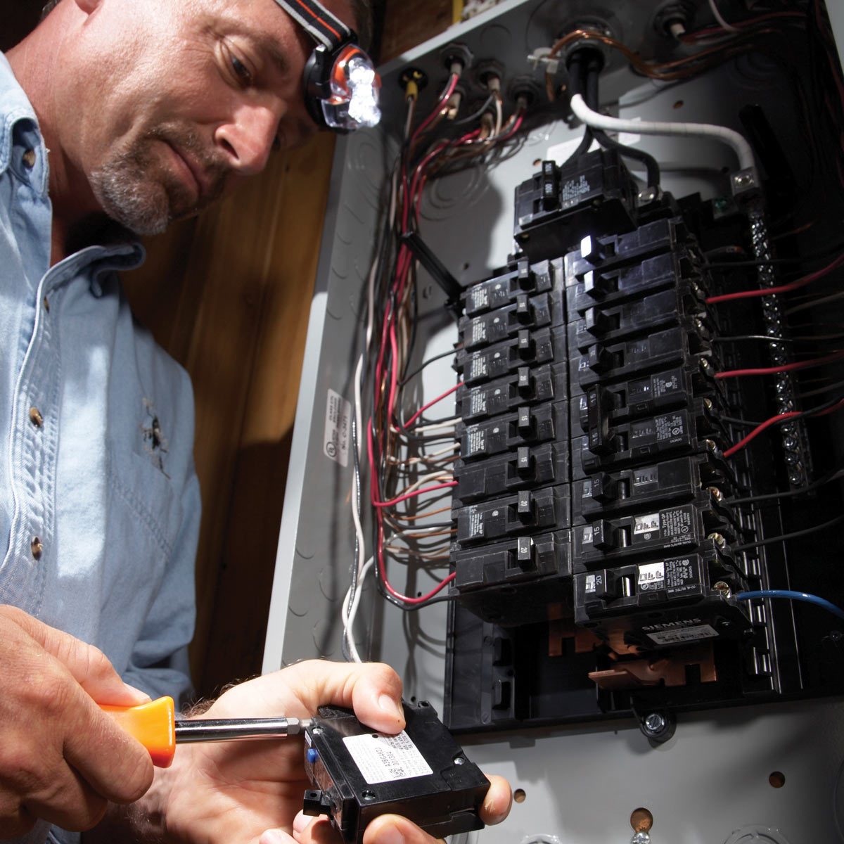 A man uses a screwdriver to replace a circuit breaker in an electrical panel, illuminated by a headlamp in a dimly lit setting.