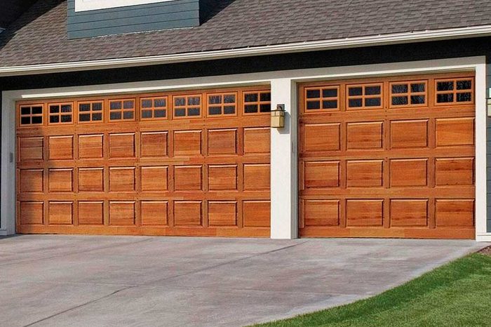 A pair of wooden garage doors are closed, flanked by light fixtures, on a house with a sloped roof under a clear blue sky and green grass nearby.