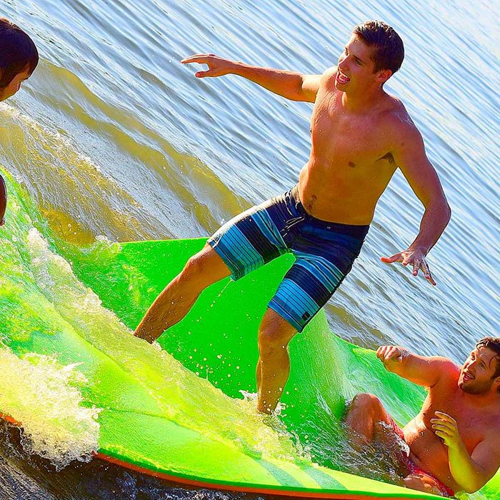 A person balances on a green surfboard, riding a wave while others splash nearby in shallow water under bright sunlight.