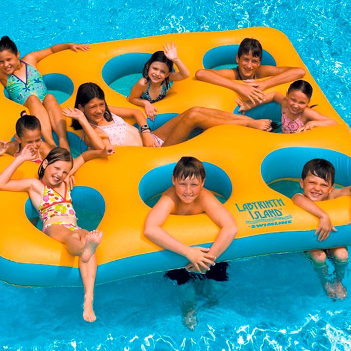 A large, yellow inflatable float with cutouts supports multiple children, who smile and wave while enjoying a sunny day in a swimming pool.
