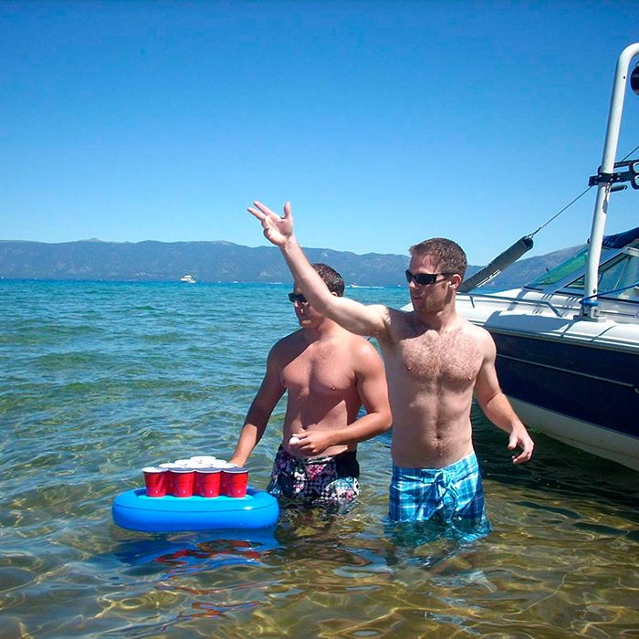 Two shirtless men stand in shallow water, one tossing a ball towards a floating beer pong setup, with a boat visible nearby and mountains in the background.