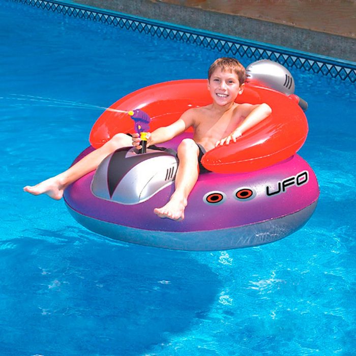 A child sits on a colorful inflatable UFO in a swimming pool, joyfully spraying water with a toy gun while smiling.