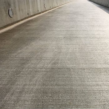 A smooth, gray concrete pathway extends forward, bordered by a vertical wall with circular openings, illuminated by natural light overhead.
