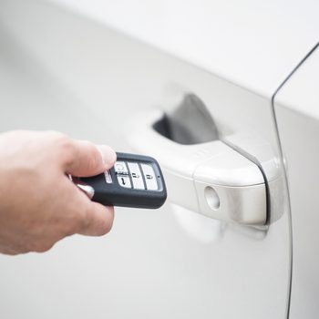 A hand holds a car key fob, pressing a button near a silver car's door handle, indicating an attempt to unlock the vehicle.