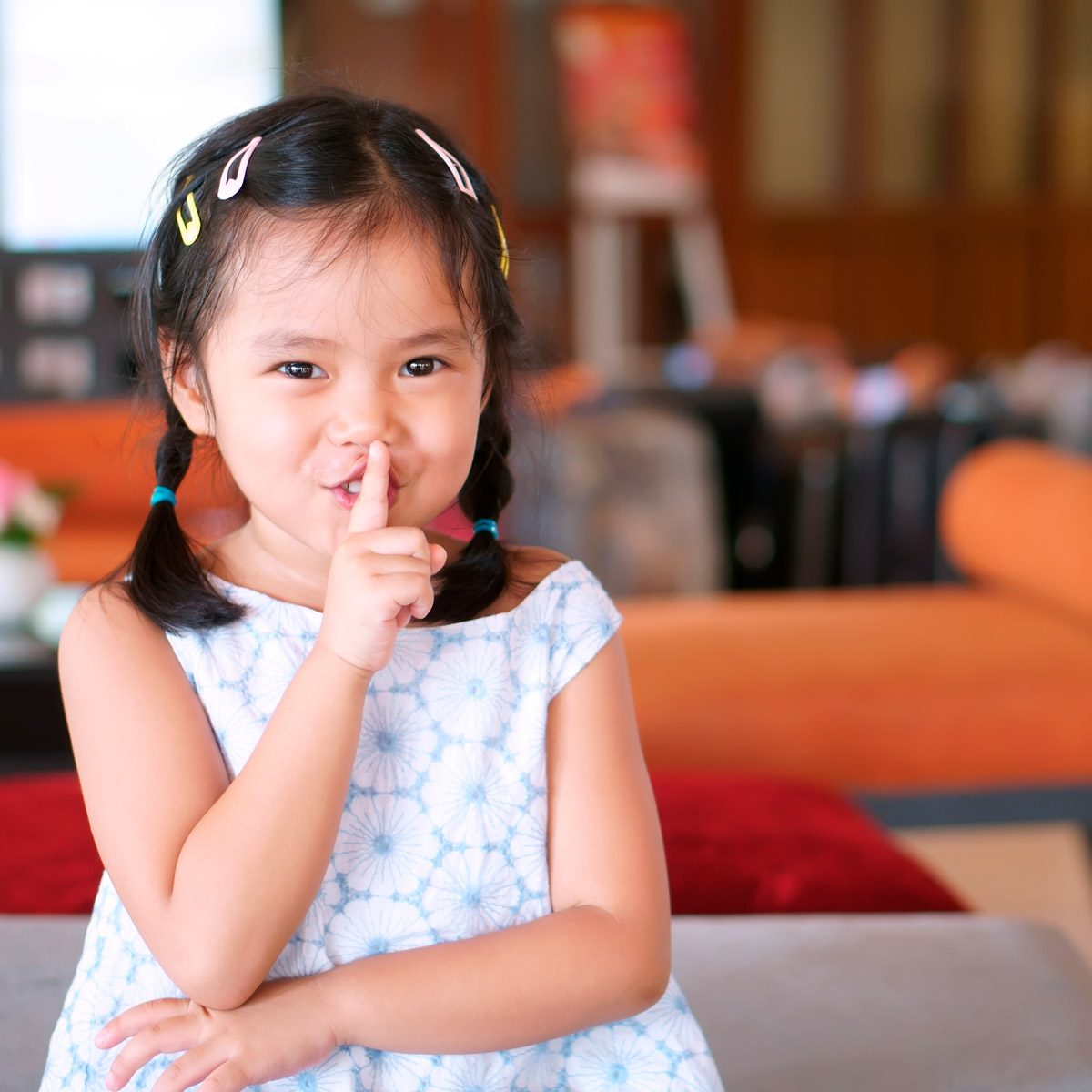 A child with pigtails playfully shushes while sitting on a couch, surrounded by a cozy indoor setting with blurred furniture in the background.