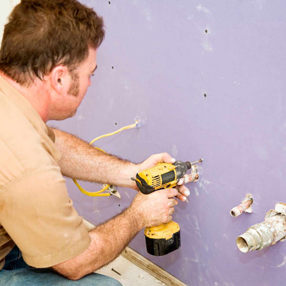 A man drills into a purple wall with a yellow power tool, surrounded by exposed electrical wires and plumbing fixtures.