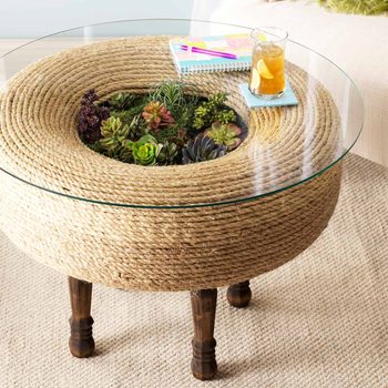 A circular table made of natural rope holds a glass top, featuring succulents inside. A drink sits on a coaster beside notebooks in a cozy room.