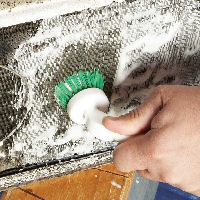 A hand holds a small scrub brush, cleaning a textured, dirty surface covered in foam, likely part of an appliance, on a wooden floor.
