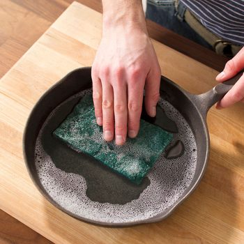 A hand scrubs a green sponge in a cast iron skillet filled with soapy water, while resting on a wooden countertop.
