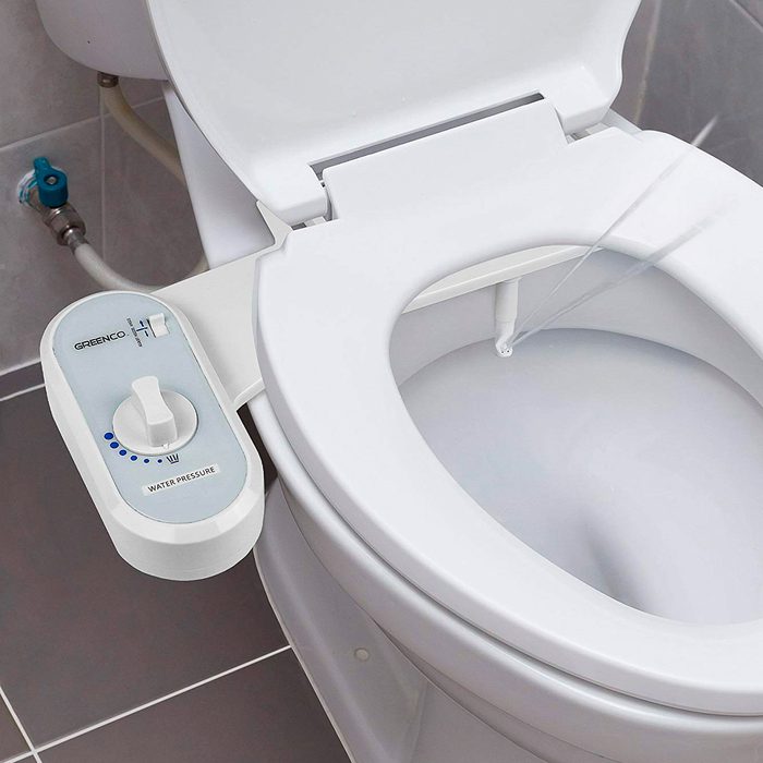 A bidet attachment, mounted on a toilet, sprays water with adjustable pressure for hygiene purposes in a bathroom setting with tiled flooring.