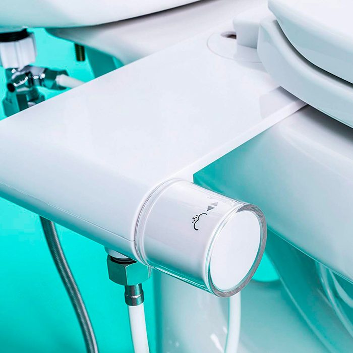 A modern bidet attachment is mounted to a toilet, featuring a control knob for water functions, set against a turquoise background.