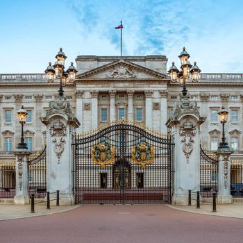 A grand wrought iron gate adorned with royal emblems opens to an ornate palace against a clear blue sky, illuminated by lamp posts lining the path.