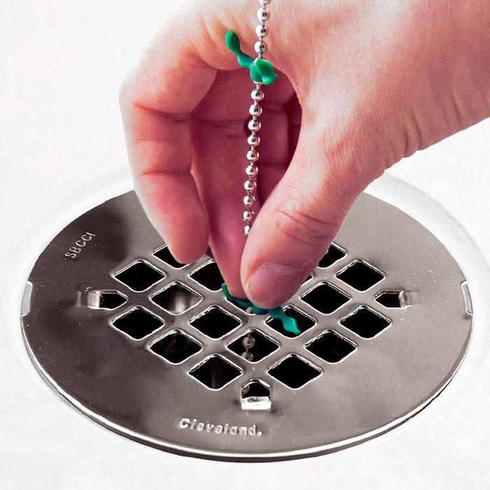 A hand holds a green object while pulling a chain from a metal drain cover with multiple openings, set against a light-colored surface.